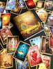 A deck of vibrant Lenormand of Enchantment cards by Schiffer Publishing, Ltd. is spread out with its guidebook, showing animals, flowers, fantasy scenes, and symbols on a dark surface.