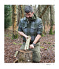 A man in green plaid kneels in a forest, splitting a log on a stump with a knife—practicing bushcraft skills just like those taught in "Core Bushcraft" by Schiffer Publishing, Ltd., for the authentic outdoor survival experience.