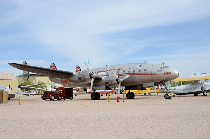 A Lockheed Constellation by Schiffer Publishing, Ltd. is displayed on a sandy lot with other planes nearby, a red support vehicle near its nose, and yellow chocks around its landing gear.