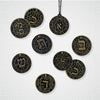 Nine bronze Hebrew Casting Lots by Schiffer Publishing, Ltd., engraved with the Hebrew Alefbet and English words like Wisdom, Justice, and Omen, are displayed on a white surface. One is shown on a black cord.