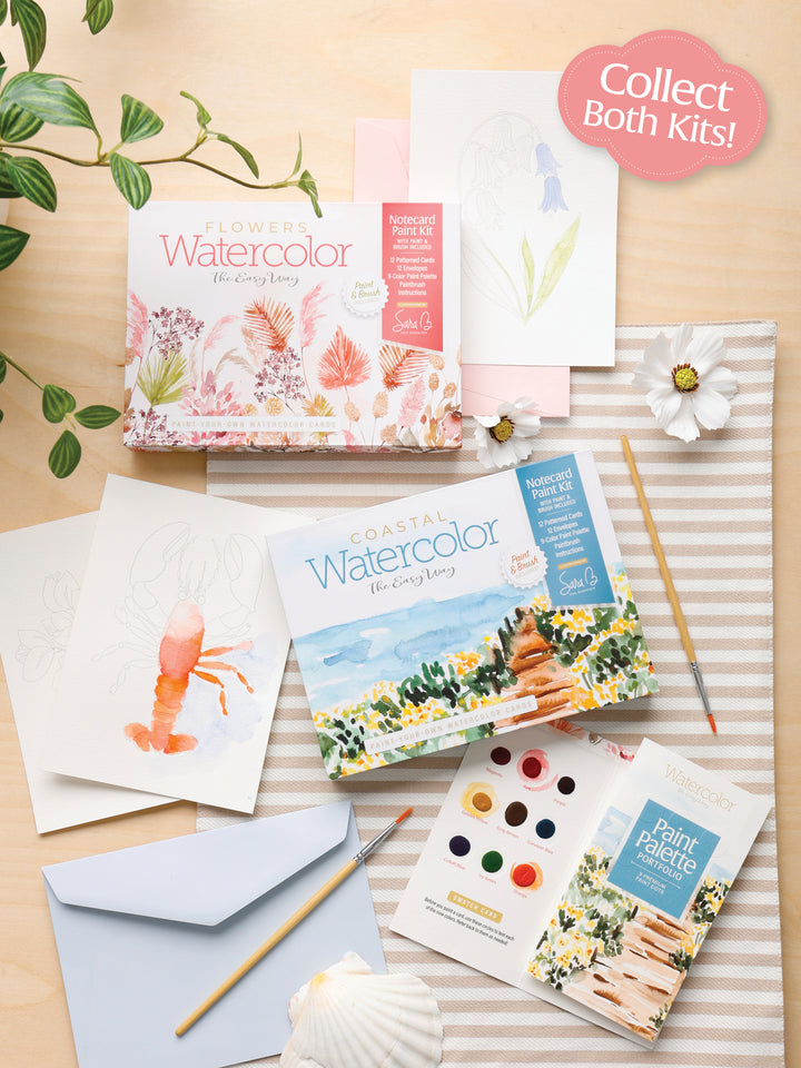 A flat lay of Schiffer Publishing, Ltd.’s Watercolor the Easy Way Paint-Your-Own Watercolor Cards Coastal kit, art supplies, floral and lobster illustrations, paint palette, brushes, a flower, envelope, notecards, and a plant on a wooden table.