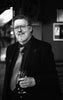 A man with glasses, beard, and mustache stands indoors in a suit and tie, holding a wine glass. The black-and-white image evokes the mysterious atmosphere of The John Dee Oracle from Schiffer Publishing, Ltd.