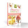 The book HeartFELT Crafts to Make and Give by Schiffer Publishing, Ltd. features colorful beginner felt projects like a frog, butterfly, fruit, and flower on its cover and stands upright against a white background.