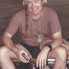 A man sits on a cot, smiling and holding a small object. Wearing a tan helmet and brown T-shirt, this Combat Historian from Schiffer Publishing, Ltd. reflects on US Army history amid his scattered belongings.