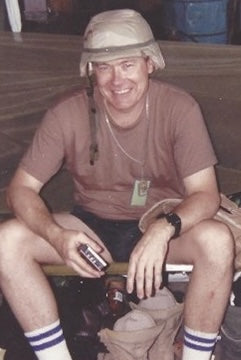 A man sits on a cot, smiling and holding a small object. Wearing a tan helmet and brown T-shirt, this Combat Historian from Schiffer Publishing, Ltd. reflects on US Army history amid his scattered belongings.