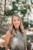 A woman with long, wavy blonde hair, styled like a pin-up, smiles outdoors in sunlit woods while wearing Dorothy's Girls by Schiffer Publishing, Ltd.—a sleeveless ribbed top and pendant necklace.