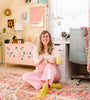 A woman with long hair and glasses, dressed in pink overalls and yellow socks, sits on a colorful rug in a pastel-themed room, smiling at the camera while holding Schiffer Publishing’s Good Vibes Art Club Learn to Draw book.