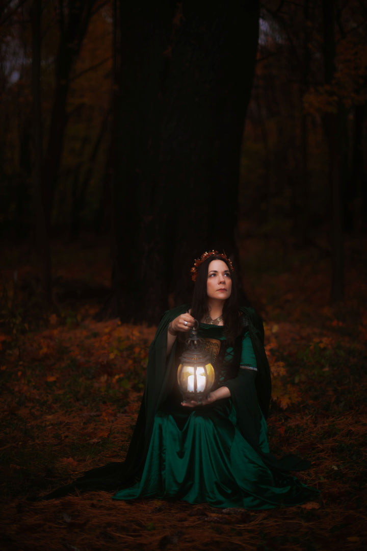 A woman in a green dress and flower crown sits on the forest floor at dusk, holding a glowing lantern amid autumn leaves and tall dark trees, evoking the rich symbolism of Schiffer Publishing, Ltd.’s The Tree Tarot.