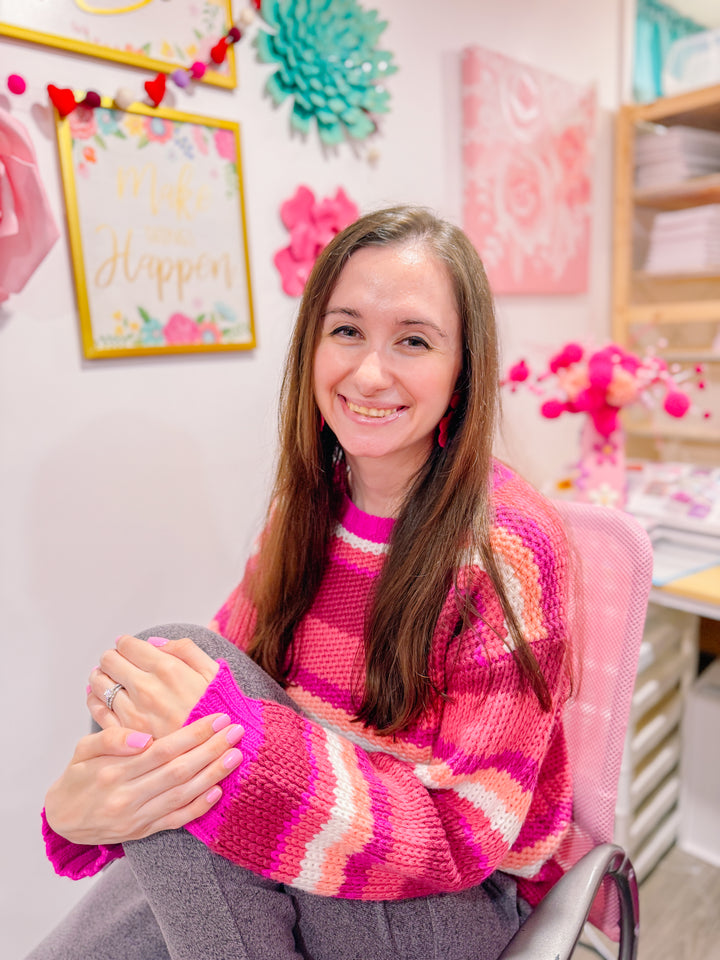 A woman with long brown hair, wearing a pink striped sweater, sits smiling in a colorful room decorated with flowers, motivational art, and adorable stickers from Schiffer Publishing, Ltd.’s *A Day in Cozy Town: The Cute Café Coloring and Sticker Book*.