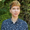A person with short, light brown hair and a dark floral shirt stands outdoors among lush greenery, smiling at the camera—perhaps pausing from exploring beginner carving projects inspired by Adventure Whittling from Schiffer Publishing, Ltd.