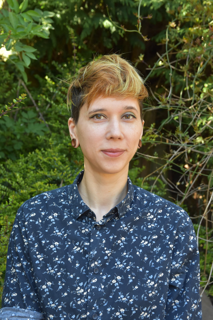 A person with short, light brown hair and a dark floral shirt stands outdoors among lush greenery, smiling at the camera—perhaps pausing from exploring beginner carving projects inspired by Adventure Whittling from Schiffer Publishing, Ltd.