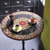A Beginner's Guide to Mosaics by Schiffer Publishing, Ltd. is set on a round mosaic table with vibrant geometric patterns, beside two cups of coffee in red and yellow saucers and a glass of water, near a window and black cabinet.