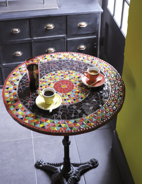 A Beginner's Guide to Mosaics by Schiffer Publishing, Ltd. is set on a round mosaic table with vibrant geometric patterns, beside two cups of coffee in red and yellow saucers and a glass of water, near a window and black cabinet.