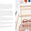 A hand uses a fork to press orange and blue yarn onto a wooden loom surrounded by labeled tools on a white surface, with instructions from Schiffer Publishing, Ltd.'s "Weaving"—ideal for learning sustainable weaving techniques.