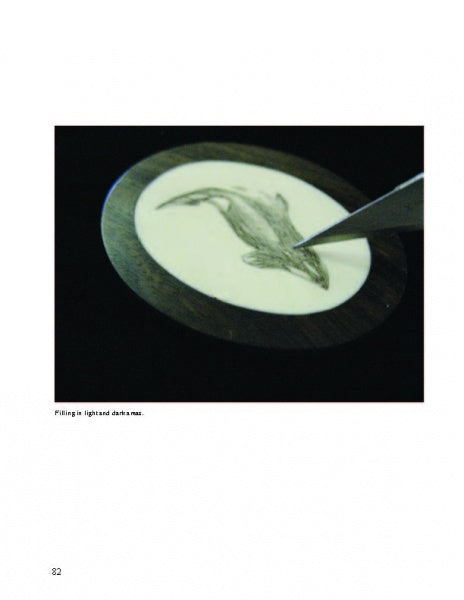 A close-up of a hand using Scrimshaw Techniques by Schiffer Publishing, Ltd. to engrave a dolphin on a round ivory substitute set in a dark frame. The tool is near the dolphin’s flipper, and the background is black.