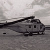 A black-and-white photo from "Desert Boneyard" by Schiffer Publishing, Ltd. shows an old helicopter without rotor blades resting in a sandy, desolate landscape beneath a partly cloudy sky—capturing retired aircraft relics in storage.
