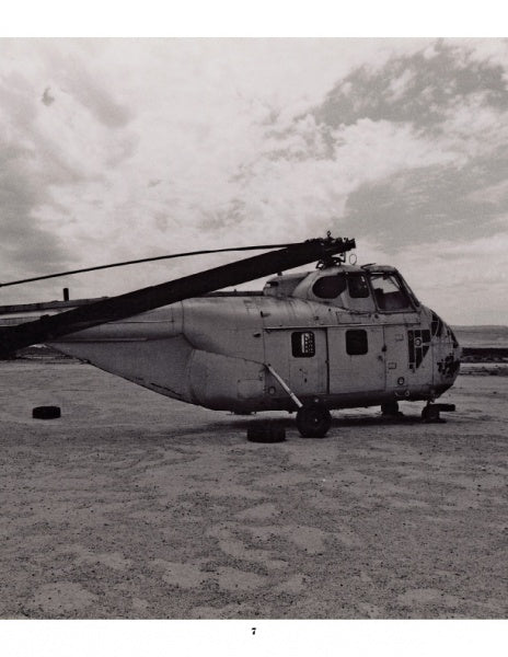 A black-and-white photo from "Desert Boneyard" by Schiffer Publishing, Ltd. shows an old helicopter without rotor blades resting in a sandy, desolate landscape beneath a partly cloudy sky—capturing retired aircraft relics in storage.