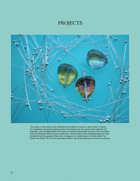 Three glass cabochons and a metal pin rest on a blue background with intersecting silver wires and beads, forming a geometric design featured in "Wire & Fused Jewelry" by Schiffer Publishing, Ltd.